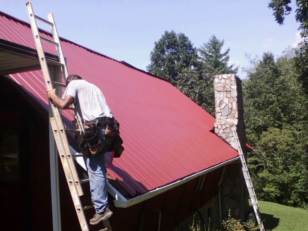 Roofer on a ladder beside a red metal roof and stone chimney — Soffit & Fascia Repair by Roofing Brothers in Thompsons Station